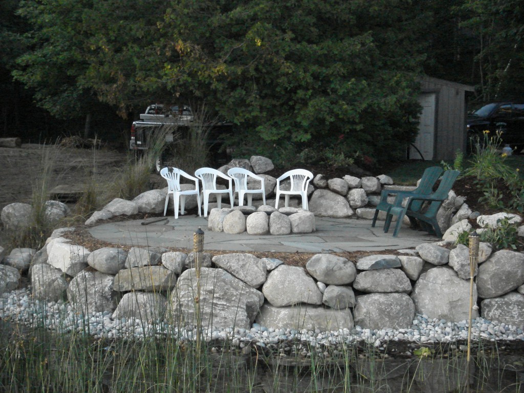 view of rocky fire pit and patio from lake